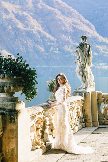 La novia con un vestido de novia se encuentra en un hermoso balcón con vistas a las montañas y al lago