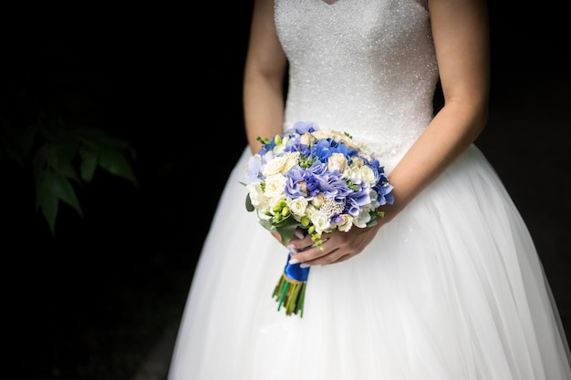 Novia en un vestido de pie en un jardín verde y sosteniendo un ramo de flores y vegetación.