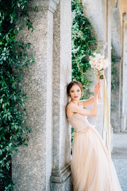 La novia con un vestido con un ramo de flores rosadas está apoyada contra un pilar con los brazos levantados en