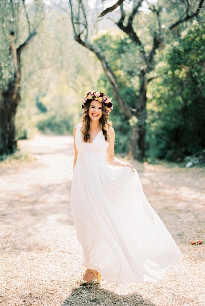 La novia se para con un vestido de toga blanca sosteniendo el dobladillo con la mano en el parque