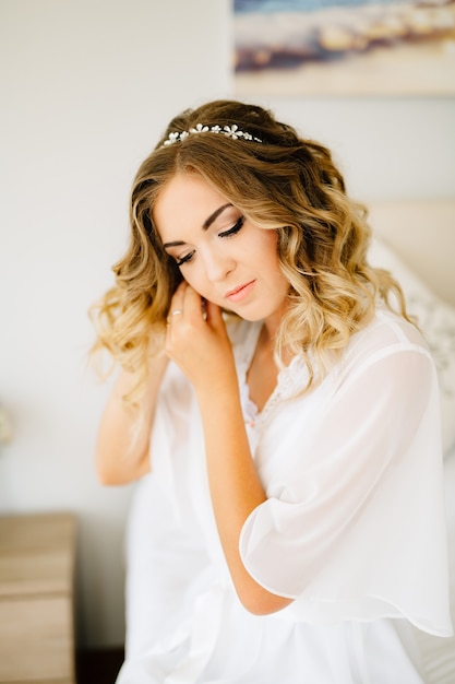 Foto novia vistiendo bata blanca se pone un pendiente mientras se prepara para la boda