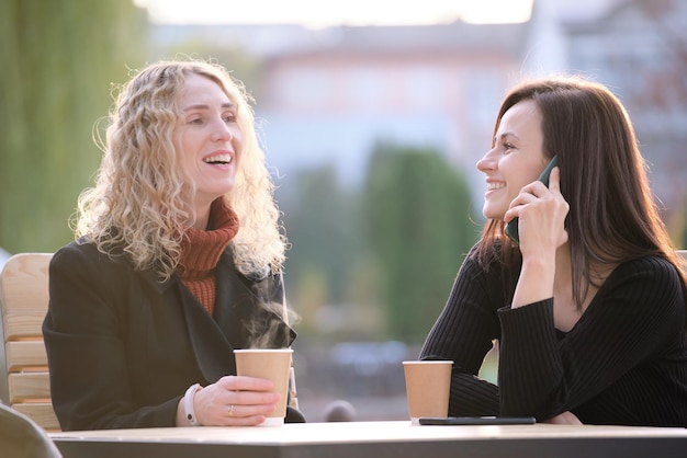 Novias felices divirtiéndose juntas sentadas en un café callejero urbano riendo y hablando por teléfono celular Concepto de amistad y actividades al aire libre