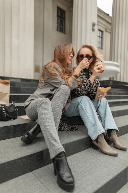 Novias de mujeres jóvenes divertidas de moda en ropa de moda con estilo se sienta en los escalones, bebe café y come profiteroles en la ciudad Las niñas felices viajan y descansan estilo de vida