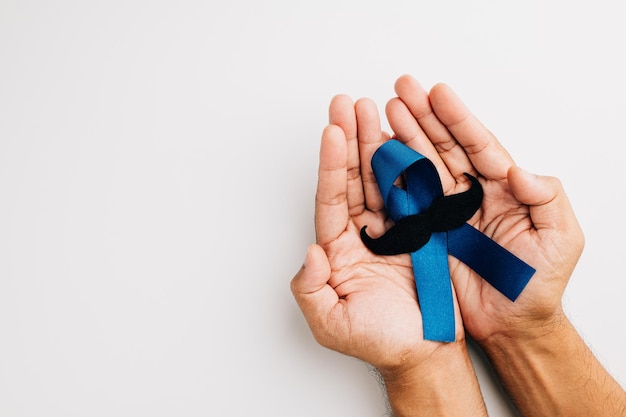 En noviembre, la salud de los hombres y la concientización sobre el cáncer de próstata toman protagonismo. Las manos del hombre abrazan una cinta azul claro con un bigote sobre un fondo azul, un símbolo de apoyo.