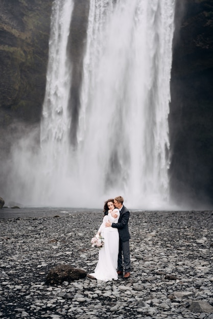 El novio abraza a la novia por detrás de la pareja de novios cerca de skogafoss cascada destino islandia