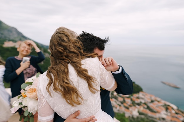 El novio abraza a la novia con un ramo de flores mientras está de pie en la vista trasera de la montaña