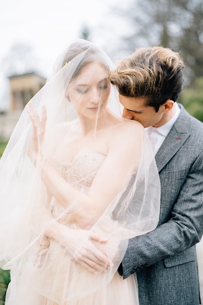 El novio abraza a la novia con un velo desde atrás y la besa en el hombro en el jardín del lago de como italia