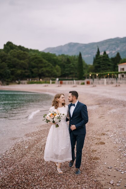 El novio abraza a la novia con un vestido blanco con un ramo de flores caminando con ella por la playa de guijarros