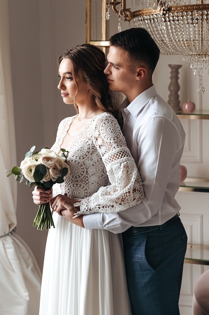 El novio abraza suavemente a la novia con un hermoso vestido de encaje con un ramo de flores frescas. Sesión de fotos de boda