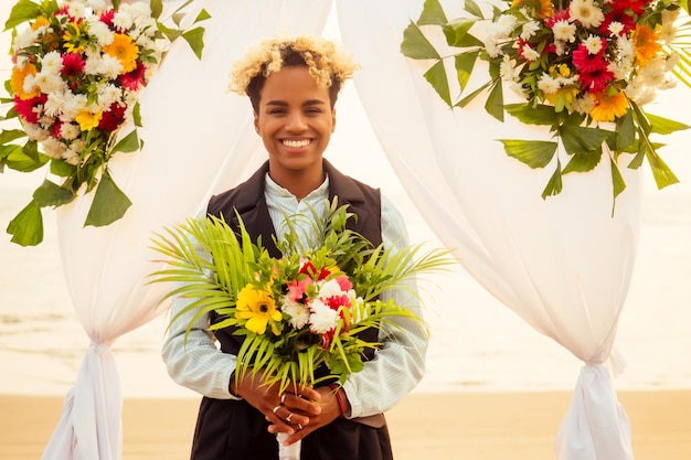 Novio afroamericano con traje negro y novia caucásica vestida en ceremonia en una playa tropical bajo el arco de la boda