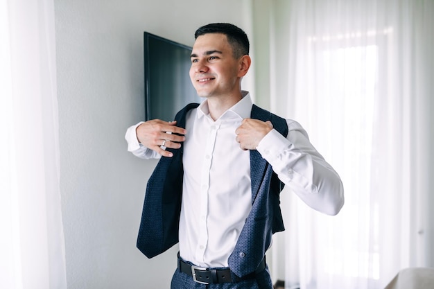 El novio alegre sonríe poniéndose un chaleco en una habitación moderna y luminosa Preparación de la boda por la mañana