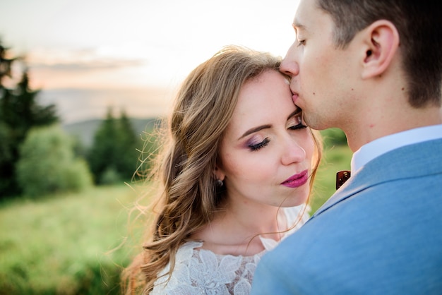 El novio besa la frente de la novia que se coloca en una colina verde