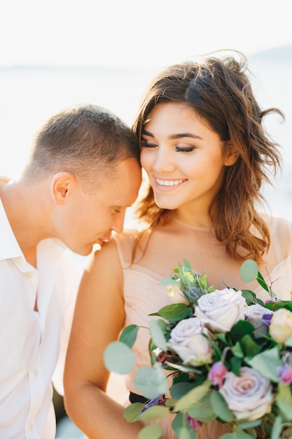 Novio con camisa blanca besa en el hombro a una novia sonriente con un hermoso vestido pastel con un