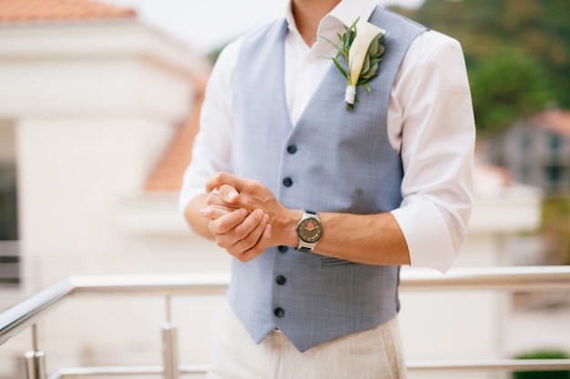 El novio con una camisa y un chaleco con un boutonniere se encuentra en el balcón en la mañana antes de la