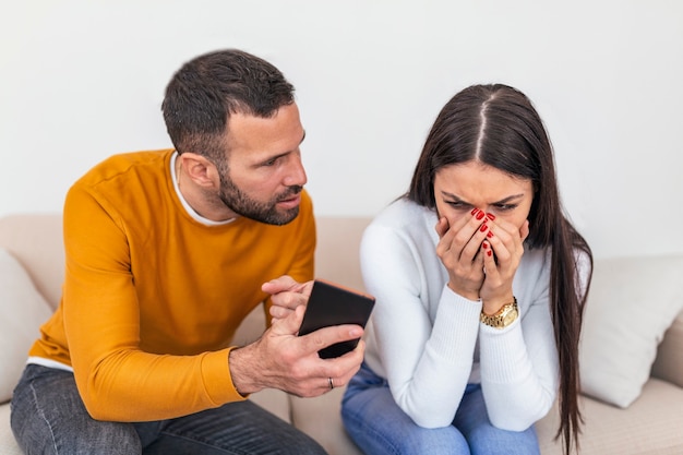 Novio celoso mostrando a su novia infiel su teléfono exigiendo explicación sentado en el sofá interior.