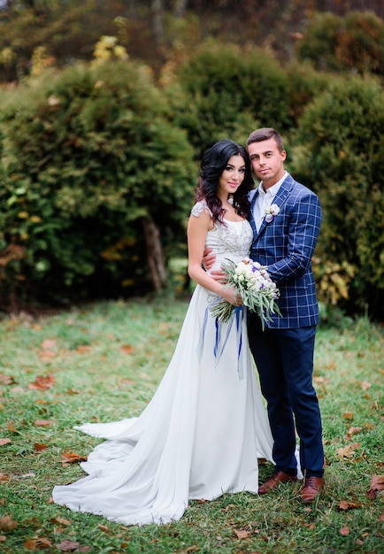 El novio en la chaqueta azul escocesa sostiene la cintura de la novia que se coloca con ella en césped verde