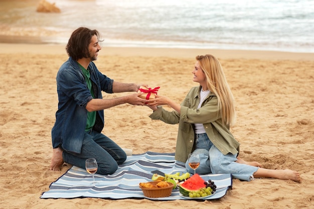 Novio dando un regalo a una novia sorprendida mientras hacen un picnic en la playa