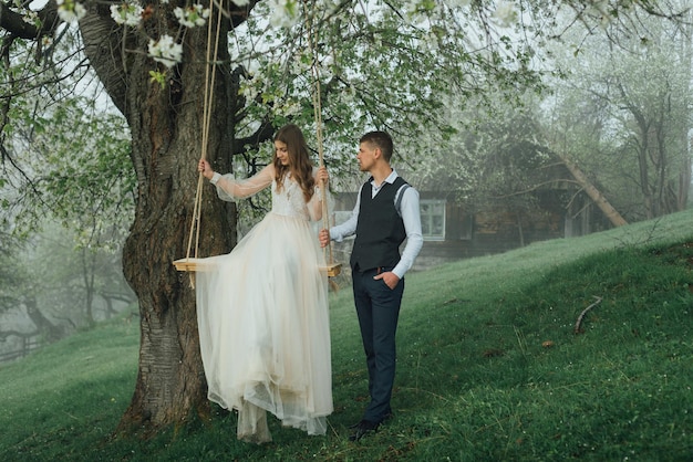 Un novio elegante hace rodar a la novia en un columpio, con el telón de fondo de la hermosa naturaleza y la hierba verde. Tiempo feliz en la boda. Graciosos recién casados están bromeando.