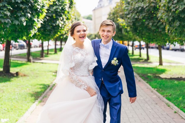 El novio con un elegante traje abraza a la novia y mira a la cámara caminar en el parque