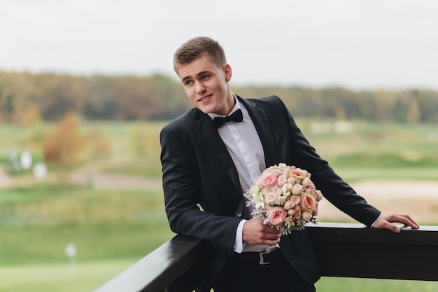 Novio esperando a la novia novio guapo al aire libre en traje negro de pie y sosteniendo ramo de novia