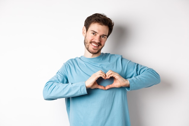 Novio guapo diciendo te amo, mostrando gesto de corazón y sonriendo a la cámara, expresa amor y sentimiento romántico, de pie sobre fondo blanco