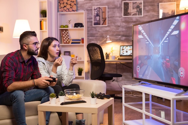 Novio jugando videojuegos en la televisión usando el controlador mientras la novia está comiendo papas fritas. Pareja sentada en el sofá.