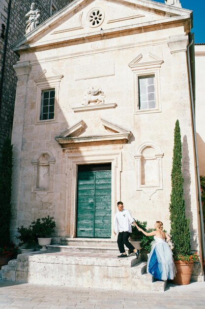 Foto el novio lleva a la novia de la mano por los escalones de la iglesia de san marcos el apóstol en perast, montenegro.