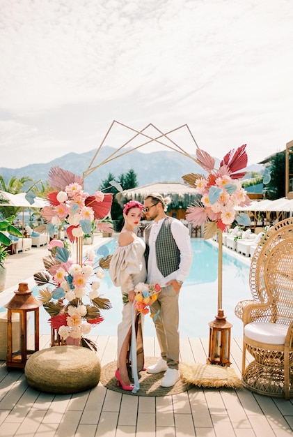 El novio mira a la novia de pie en el arco colorido de la boda en la orilla de la piscina