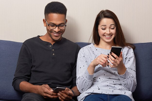 El novio y la novia adictos a la raza mixta contentos tienen expresiones faciales felices, tienen teléfonos inteligentes modernos