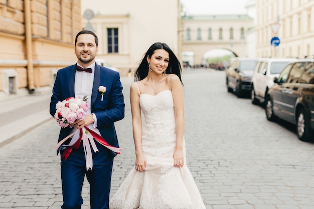 El novio y la novia caminan juntos, posan ante la cámara, tienen expresiones complacidas, celebran su boda