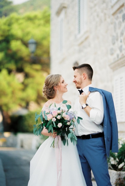 El novio y la novia con un ramo de flores se paran cerca del edificio de piedra