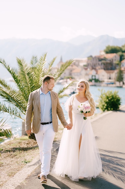 El novio y la novia sonriente con un ramo caminan tomados de la mano en el muelle cerca de la palmera