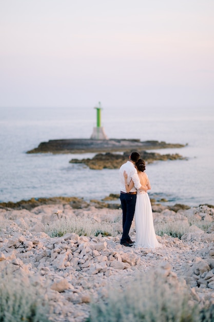 El novio y la novia con un vestido blanco están de pie, abrazados, en el cabo de Punta Planca, admirando el faro verde en el mar en un día soleado. Foto de alta calidad