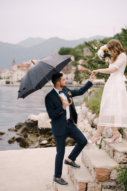 El novio bajo el paraguas ayuda a la novia a bajar los escalones del muelle.