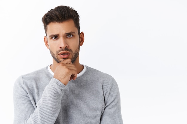 Foto un novio preocupado de aspecto serio que escucha a un amigo tiene un problema, tratando de averiguar cómo ayudar, de pie en la pared blanca, tocando la barbilla pensativo, frunciendo el ceño mientras escucha atentamente la conversación