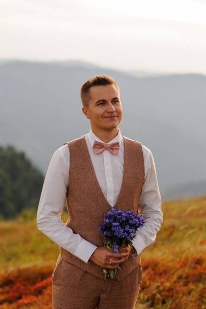 Un novio con un ramo de flores silvestres mira a su novia. Fotografía de bodas en las montañas.