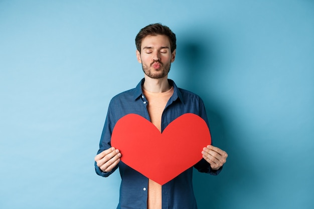 Novio romántico con ojos cerrados de corazón rojo de San Valentín, labios fruncidos y esperando un beso en el día de los enamorados, de pie contra el fondo azul.