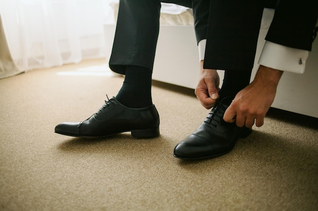 El novio sentado en el sofá y atado los cordones de los zapatos de cuero negro, primer plano. boda