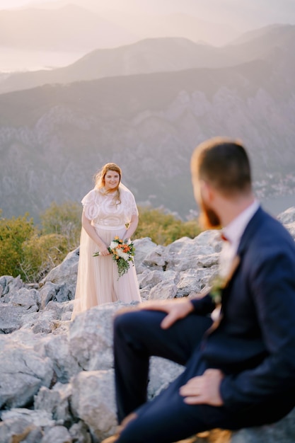 El novio se sienta sobre las piedras y mira a la novia de pie con un ramo