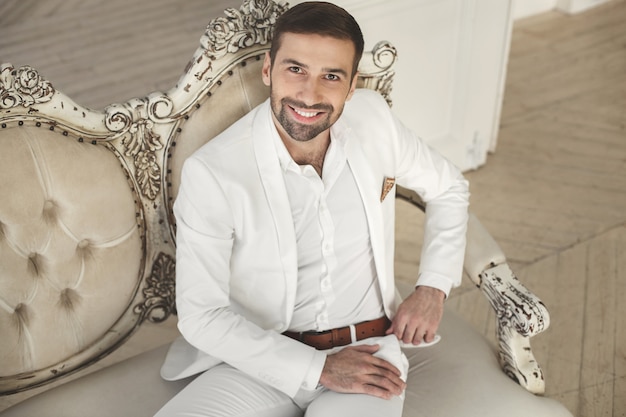 El novio sonriendo y mirando a la cámara. Elegante joven guapo con barba en un traje clásico blanco.