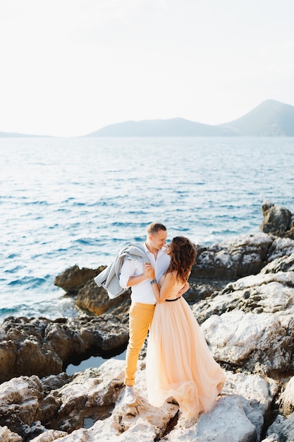 Novio sonriente abraza a la novia en un hermoso vestido beige de pie sobre una roca