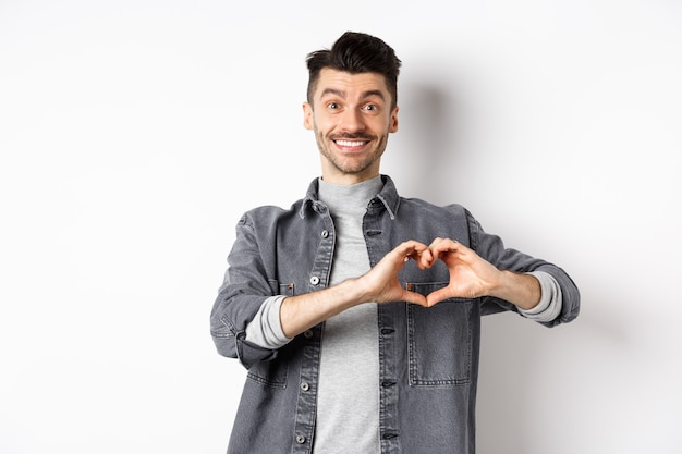 Novio sonriente mostrando corazón con amor, mirando romántico y feliz al amante, de pie sobre fondo blanco. Concepto de relación y día de San Valentín.