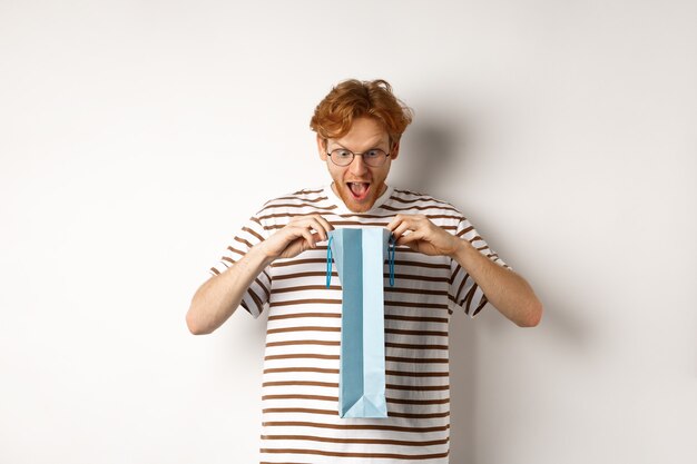 Novio sorprendido abra la bolsa de compras con el regalo del día de San Valentín, mirando hacia adentro con cara de asombro, de pie sobre fondo blanco.