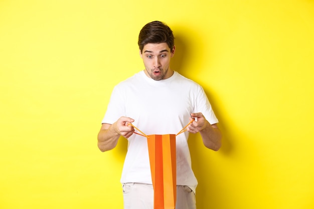 Novio sorprendido bolsa abierta con regalo, mirando dentro asombrado, de pie sobre la pared amarilla