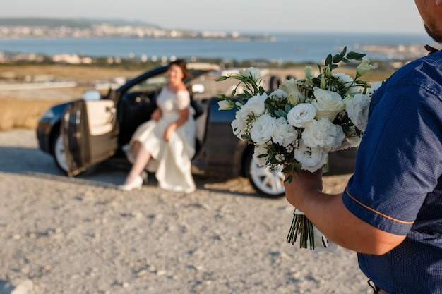 Novio sosteniendo flores de ramo de boda y ve al puente sentado en el coche de bodas