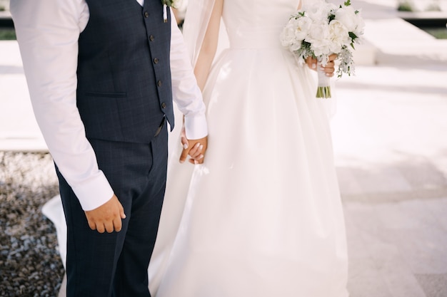 El novio sostiene la mano de la novia con un vestido blanco con un ramo de flores