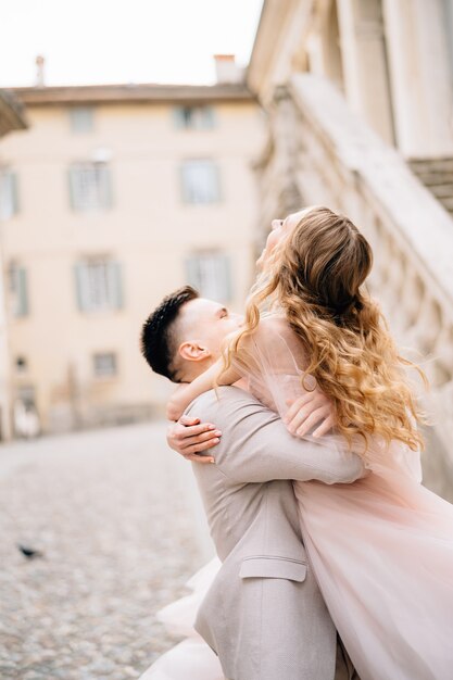 El novio sostiene a la novia en sus brazos contra la escena de un edificio antiguo en Bérgamo Italia