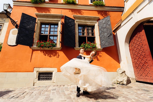 El novio sostiene a su novia en brazos y bailan cerca del edificio con hermosas ventanas.