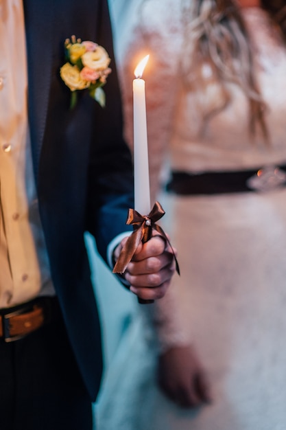 El novio sostiene una vela de boda con un lazo marrón.