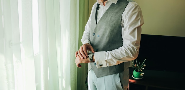 El novio con traje gris claro está parado cerca de la ventana Hombre mirando su reloj Ropa elegante